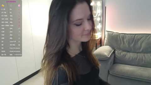 Media: Video of a young woman with long, straight brown hair, wearing a black top, standing indoors in a dimly lit living room with a gray couch and a lamp.