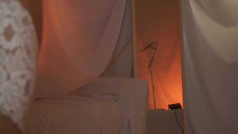 Media: A video depicts a dimly lit, cozy bedroom scene with a white bed and a small lamp emitting a warm orange glow, creating a soft, intimate ambiance.