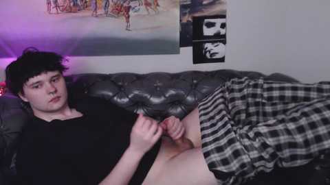 Media: A video of a young white man with short black hair, wearing a black shirt and plaid pants, lying on a tufted leather couch. The background includes a wall with posters and a purple light.