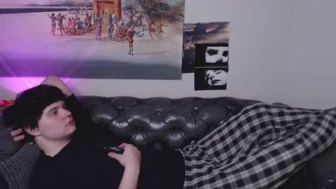 Media: Video of a young person with short black hair, wearing a black shirt, lying on a dark gray leather couch with a black and white checkered blanket, holding a controller, in a room with posters and a purple light.