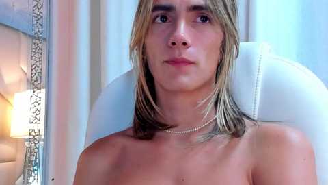 Media: Video of a topless, fair-skinned woman with long, straight, blonde hair, wearing a pearl necklace, seated on a white leather chair in a softly lit room with ornate silver wallpaper.