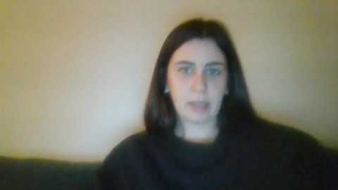 Media: A blurry video of a young woman with long, dark hair, wearing a black top, against a plain beige wall. Her facial expression is indiscernible due to the low resolution and blur.