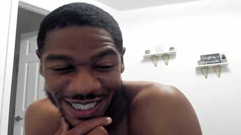 Media: Video of a shirtless, muscular Black man with a trimmed beard and braces, smiling broadly, in a modern, white-walled bathroom with wooden shelves and a door visible in the background.