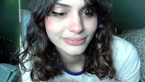 Media: Video of a young woman with curly brown hair, light skin, and rosy cheeks, wearing a white t-shirt with blue trim, smiling in a dimly lit room.