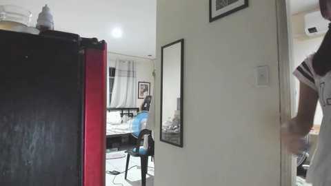 Media: Video of a man in a white shirt standing in a doorway, looking into a brightly lit room with a desk, chair, and framed artwork on beige walls.