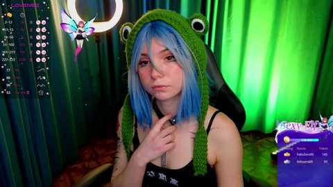 Media: Video of a pale-skinned woman with blue hair in a green frog hat, wearing a black tank top, in a dimly lit room with green and blue lights, holding a necklace.