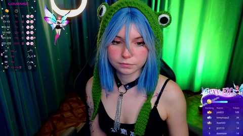 Media: Video of a young woman with blue hair, wearing a green frog hat, black tank top, and a choker, sitting in a dimly lit room with green and purple lights.