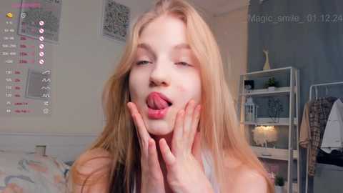 Media: Video of a blonde teenage girl with fair skin, wearing a white tank top, sticking out her tongue in a playful manner. The background shows a modern, lightly decorated bedroom with a calendar, bookshelf, and a bed.