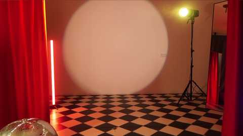 Media: Video of a dimly lit, sparsely furnished room with a large, circular spotlight casting a bright yellow glow on a white wall. The floor has a black-and-white checkered pattern, and red curtains frame the scene.