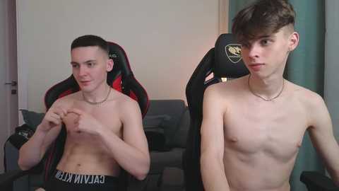 Media: Video of two shirtless young men with pale skin, one with short dark hair, the other with a buzz cut, sitting in gaming chairs in a modern living room.