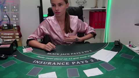 Media: Video of a light-skinned woman with long brown hair, wearing a pink satin robe, sitting at a blackjack table in a casino, surrounded by chips and cards.