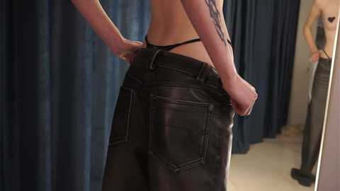 Media: Video of a slender, fair-skinned woman with a tattoo on her arm, wearing black leather pants and a black thong, standing in front of a mirror, reflecting her profile and a dark curtain in the background.