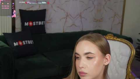 Media: Video of a young woman with fair skin, straight brown hair, and light makeup sitting on a green velvet couch with a white, gold-framed chair in the background. She has a neutral expression.