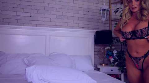 Media: Video of a blonde woman in black lace lingerie standing beside a white bed with white pillows in a dimly lit, cozy bedroom with white brick walls and a nightstand with flowers.