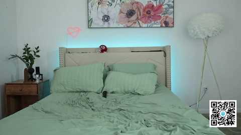 Media: Video of a neatly made bed with light green bedding, against a white wall with a floral painting and a pink heart sticker. A white floor lamp and wooden nightstand with a vase and books are visible.