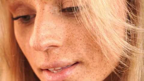 Media: Video of a close-up of a fair-skinned woman with long blonde hair, showing her freckled face, with a soft smile. The background is out of focus, emphasizing her natural beauty and gentle expression.