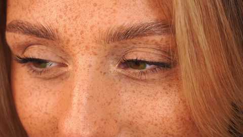 Media: Close-up video of a woman's face, showing her freckled skin, green eyes, and long blonde hair, with a blurred background.