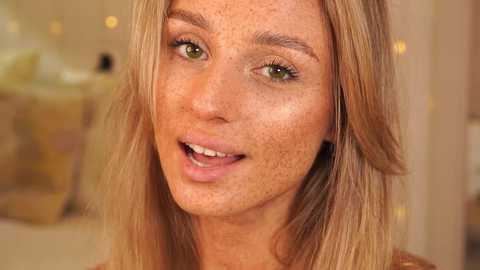 Media: Video of a fair-skinned woman with long, straight blonde hair and freckles, smiling widely, with green eyes. The background is blurred, featuring soft, warm lighting and decorative elements.