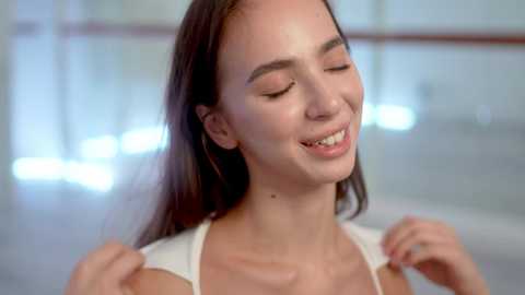 Media: Video of a young, fair-skinned woman with long, dark brown hair, wearing a white top, smiling and closing her eyes, with a blurred, modern indoor background.