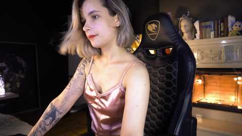 Media: Video of a fair-skinned woman with short blonde hair in a pink satin camisole, sitting in a black gaming chair with a Batman logo. Background features a lit fireplace and a bookshelf.