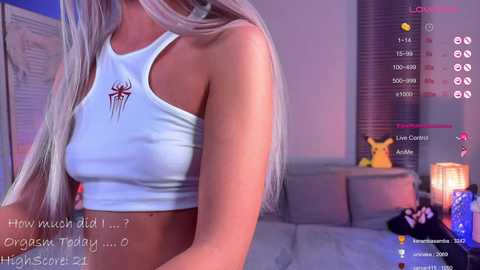 A video of a young woman with long, straight, platinum blonde hair, wearing a white crop top with a spider web design. The background features a cozy, dimly-lit living room with Pikachu plushies and a lamp.