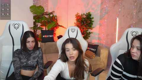 Video of three women in white gaming chairs with skull logos, wearing casual clothing, in a modern, dimly lit room with green plants, a black couch, and neon lights.