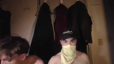 Media: Video of a shirtless young man wearing a yellow mask, standing in a dimly lit room with a wall of dark coats.