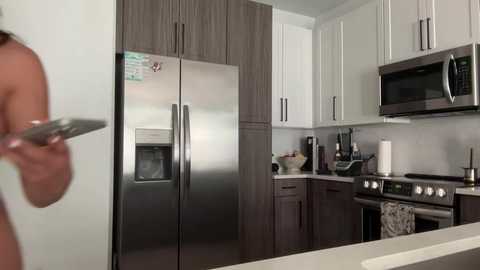 Media: Video of a modern, stainless steel double-door fridge in a sleek, minimalist kitchen with white cabinets and dark wood accents. A person, blurred, holds a knife on the left.