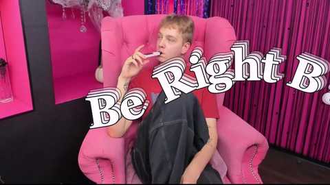 Media: Video of a young, slender, light-skinned man with short, blonde hair, wearing a red T-shirt, sitting in a pink, tufted armchair, licking a lollipop. Background is pink and black with \"Be Right B\" text.