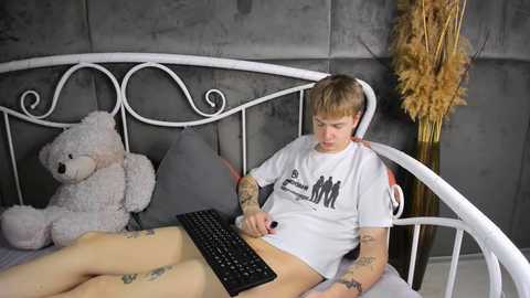 Media: A video of a tattooed man with short hair, wearing a white T-shirt, sitting on a bed, typing on a keyboard, surrounded by a gray wall and a beige teddy bear.