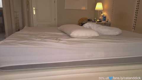 Media: Video of a neatly made bed with white linens in a softly lit, beige-walled bedroom. The room features a nightstand with a lamp, a bottle, and a framed picture.