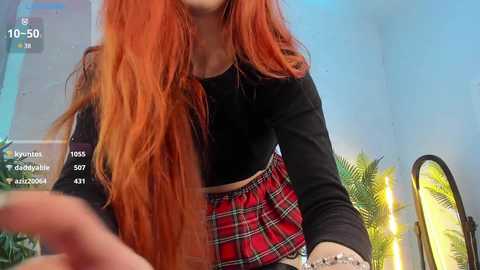 Media: Video of a young woman with long, vibrant orange hair, wearing a black crop top and a red plaid skirt. The background features a palm tree and a blue wall.