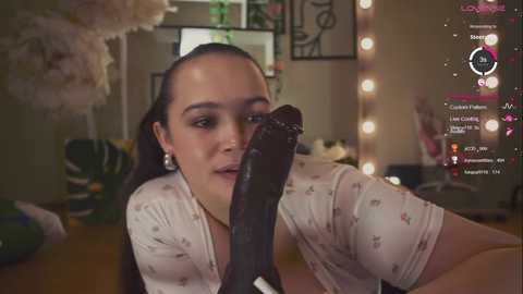 Media: Video of a young, light-skinned woman with dark hair, wearing a white floral shirt, licking a large, dark dildo, in a dimly lit bedroom with a pink light and gaming interface overlay.