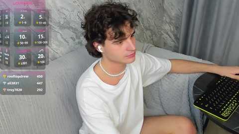 Media: Video of a young man with curly brown hair, wearing a white t-shirt, sitting on a grey couch, playing a black keyboard with yellow accents.