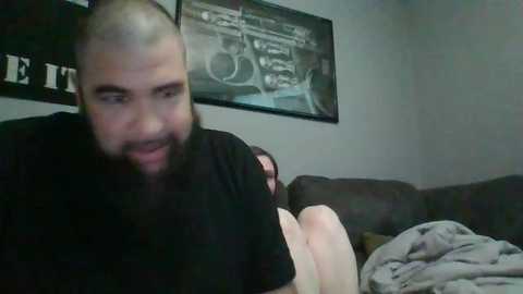 Media: Video of a bald, bearded man with a light beard in a black T-shirt, sitting on a couch next to a woman with long brown hair, both watching a movie.