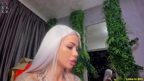 Media: Video of a woman with platinum blonde hair, wearing a lace top, standing in a room with dark curtains, green ivy walls, and a mirror.