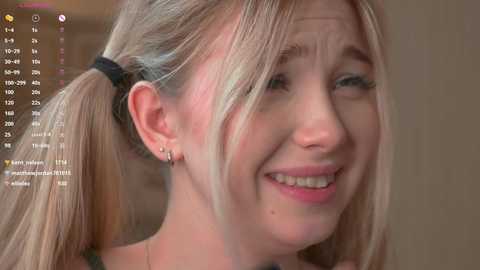 Media: Video of a smiling, fair-skinned woman with blonde pigtails, wearing a black headband, and small earrings. She has a round face, light makeup, and is seen from the shoulders up against a blurred, neutral background.