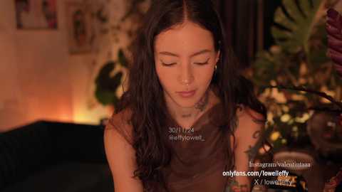 Media: Video of a young woman with long black hair, wearing a brown sleeveless top, sitting in a cozy room with warm lighting, surrounded by plants and a fireplace.