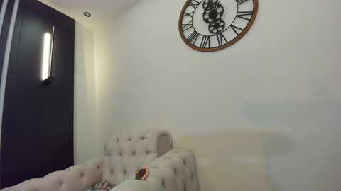 Media: A video of a minimalist living room with a beige tufted sofa, a large white wall clock, and a dark wooden door.