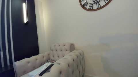 Media: A video of a modern living room corner, featuring a tufted beige sofa with a black remote control on it, a wall clock, and a vertical strip of white lights on a dark wall.