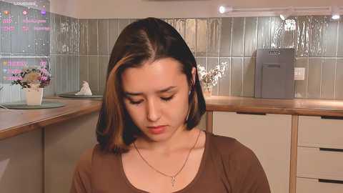 Media: Video of a young woman with straight, shoulder-length brown hair, wearing a brown top, seated in a modern kitchen with tiled backsplash and wooden countertops.