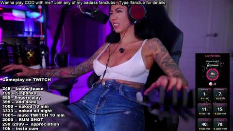 Media: Video of a tattooed woman with large breasts, wearing a white crop top and high-waisted jeans, playing video games in a dimly lit room with a gaming chair and a computer monitor.