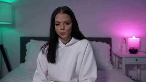 Media: Video of a young woman with straight black hair, wearing a white bathrobe, sitting on a bed with white sheets in a dimly lit bedroom. A nightstand with a lit candle and a lamp with green and pink lights is visible.