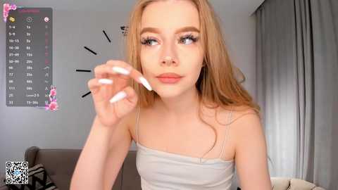 Media: Video of a young, light-skinned woman with long blonde hair, wearing a white tank top, applying makeup with long, white nails. Background shows a calendar and curtains in a softly lit room.