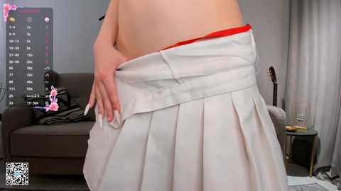 Media: Video of a woman's bare back, lifting a pleated white skirt, revealing a red thong. Background includes a gray couch, calendar, guitar, and a glass of wine.