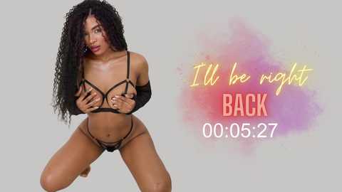 Media: Video of a dark-skinned woman with voluminous curly black hair, wearing black strappy lingerie and elbow-length gloves, kneeling provocatively against a plain white background. Text overlay reads: \"I'll be right BACK 00:05:27.\