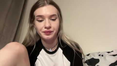 Media: A video of a young woman with fair skin, long blonde hair, and a white-and-black graphic tee. She has a subtle smile, wearing minimal makeup. Background shows a plain wall and a partly visible white and black patterned fabric.