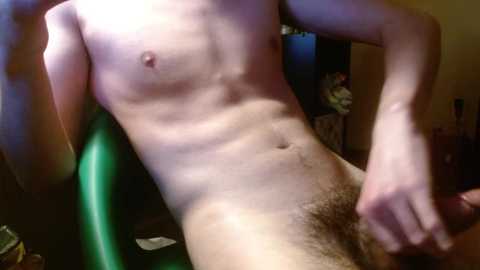 Media: Video of a nude, fair-skinned, lean male with a small penis and pubic hair, sitting on a green chair, with a blurred background showing a cluttered room.