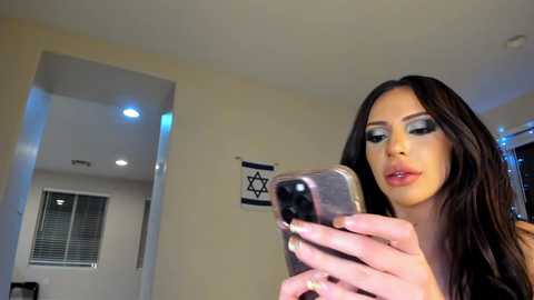 Media: Video of a light-skinned woman with long brown hair, wearing heavy makeup with blue eyeshadow, taking a selfie in a modern, minimalist room with beige walls and a Star of David on the wall.