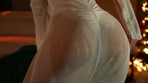 Media: Video of a woman in a sheer, white, long-sleeved dress, emphasizing her round, bare buttocks. Background features a warm, blurred Christmas tree with multicolored lights and a dark green couch.